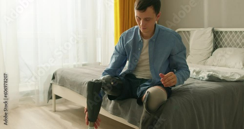 active ambitious disabled man putting on trainers, sneakers going to go in for sport Slow motion young man leg amputee wearing or changing prostheses at home sitting on bed in bright room, alone.