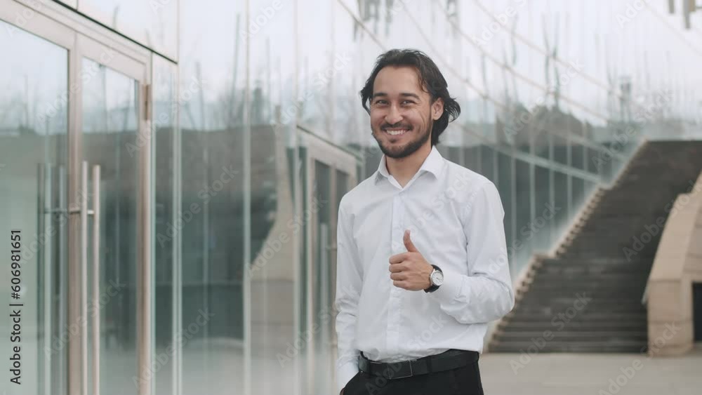 Portrait of a young happy asian businessman in a white shirt standing in front of a business center, looking with confidence, smiling and gives a thumbs up Portrait of a young happy asian businessman in a white shirt standing in front of a business center, looking with confidence, smiling and gives a thumbs up