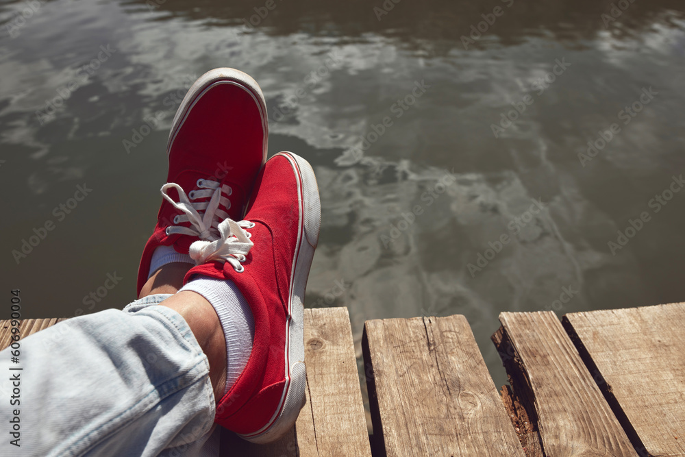 Person in red shoes relaxing on a lake wooden deck. - obrazy, fototapety, plakaty 