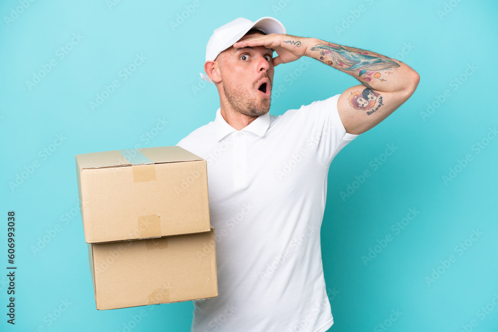 Delivery caucasian man isolated on blue background doing surprise gesture while looking to the side Delivery caucasian man isolated on blue background doing surprise gesture while looking to the side