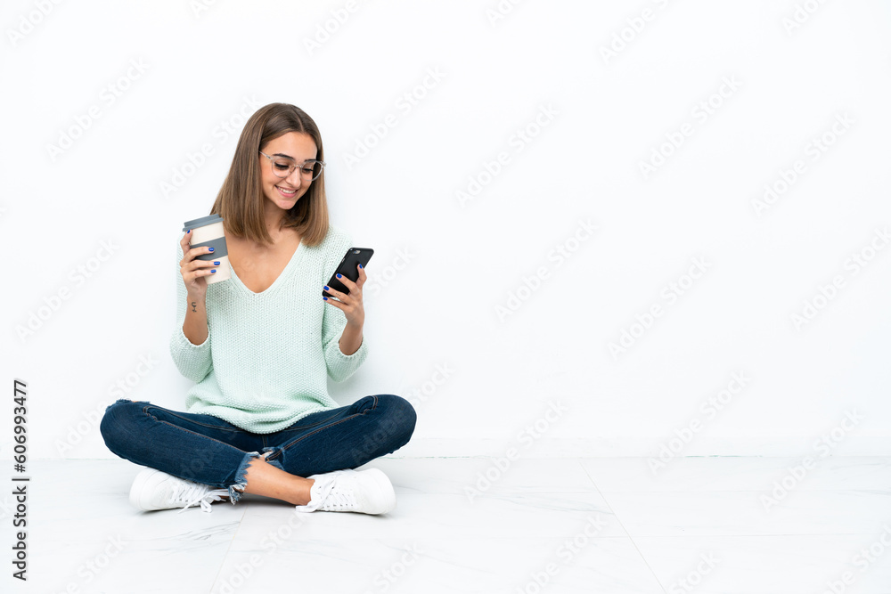 Young caucasian woman sitting on the floor isolated on white background holding coffee to take away and a mobile Young caucasian woman sitting on the floor isolated on white background holding coffee to take away and a mobile