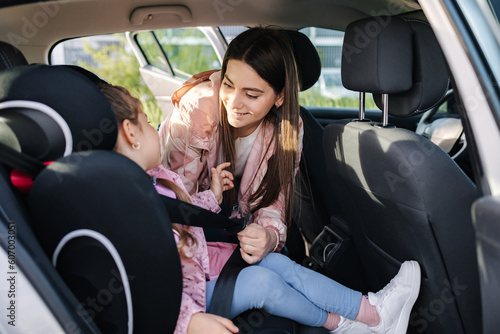 Attractive young mother put her daughter in a car seat and fastens her seat belts. Woman care about kid. Protection during the trip in the car.