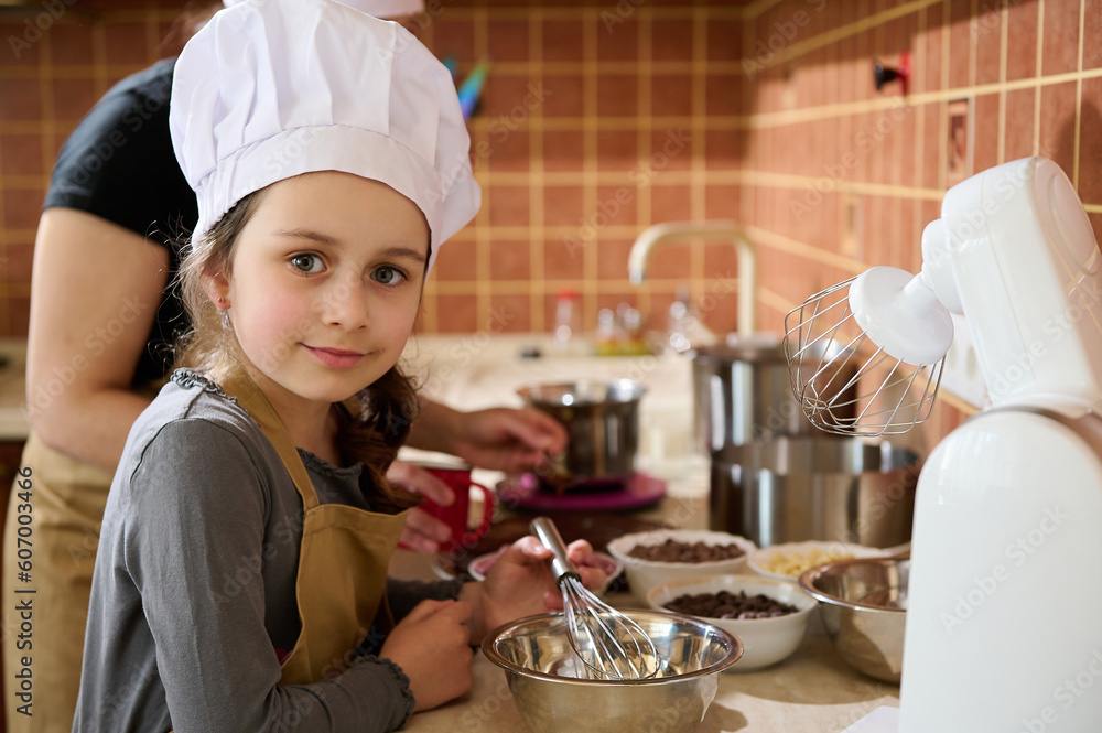 Caucasian little girl in chef's hat and kitchen apron, smiling, looking at camera, helping her mother to prepare a delicious homemade dessert for festive occasion. mom and daughter cooking together Caucasian little girl in chef's hat and kitchen apron, smiling, looking at camera, helping her mother to prepare a delicious homemade dessert for festive occasion. mom and daughter cooking together