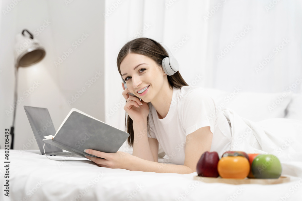 woman in the white shirt and pajama on a bed looking reading a book and wearing white headphones. woman in the white shirt and pajama on a bed looking reading a book and wearing white headphones.