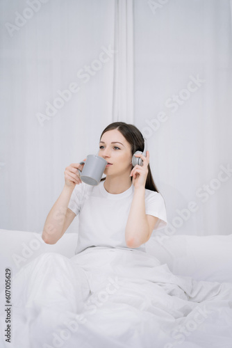 Young woman in the white shirt and pajama relaxing lying on a bed, she is listen to music and enjoys a coffee in the morning.