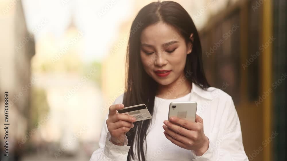 Portrait of charming young asian woman paying with credit card on smartphone at city street Happy customer doing payments online shopping in internet store and receiving cash back outdoors Portrait of charming young asian woman paying with credit card on smartphone at city street Happy customer doing payments online shopping in internet store and receiving cash back outdoors