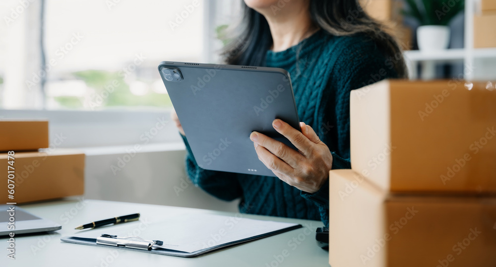 Startup small business entrepreneur or freelance using a tablet with box, checking orders from customers, woman runs an e-commerce business on websites and social media. Startup small business entrepreneur or freelance using a tablet with box, checking orders from customers, woman runs an e-commerce business on websites and social media.