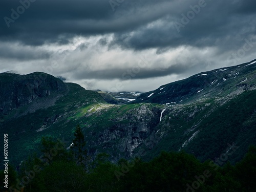 Snowy mountain top, covered with green grass, and green trees, with a stormy sky in the background