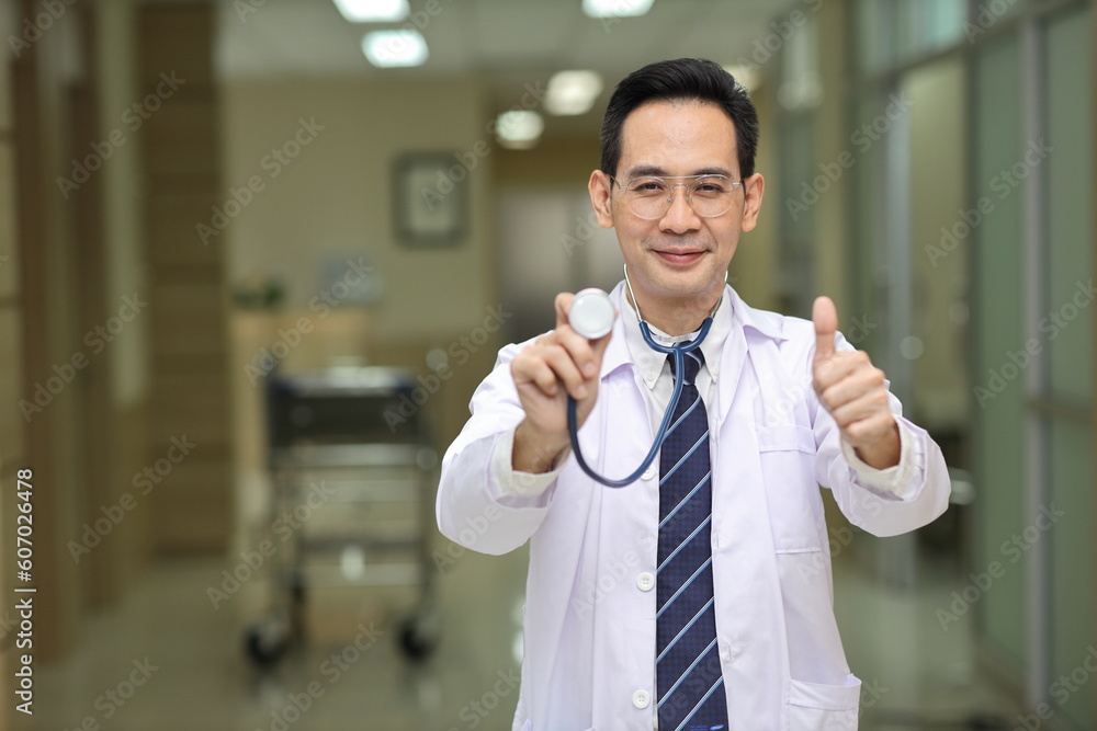 Happy asian medicine doctor standing and using stethoscope while showing thumb up and smile with hospital blurred background. Medical technology, treatment, healthcare and health insurance concept Happy asian medicine doctor standing and using stethoscope while showing thumb up and smile with hospital blurred background. Medical technology, treatment, healthcare and health insurance concept