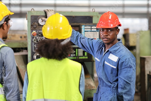 Technician engineer in protective uniform with hardhat standing and teaching apprentices or colleague worker to use computerized machine control at heavy industry manufacturing factory