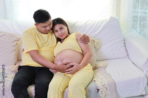 Happy asian adult couple with husband sitting and resting on sofa in living room while holding baby inside a pregnant and smile. Expectant mother preparing and waiting for baby birth during pregnancy.