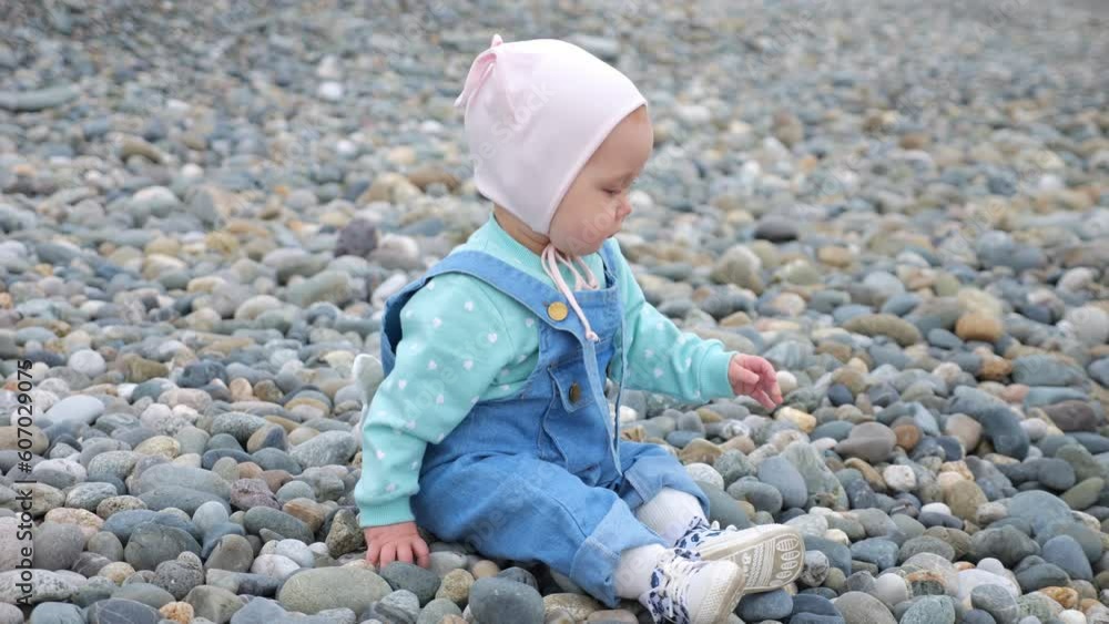 Toddler girl enjoys sitting and playing with pebbles on empty beach. Infant wearing pink hat plays with excited and delighted expression Toddler girl enjoys sitting and playing with pebbles on empty beach. Infant wearing pink hat plays with excited and delighted expression
