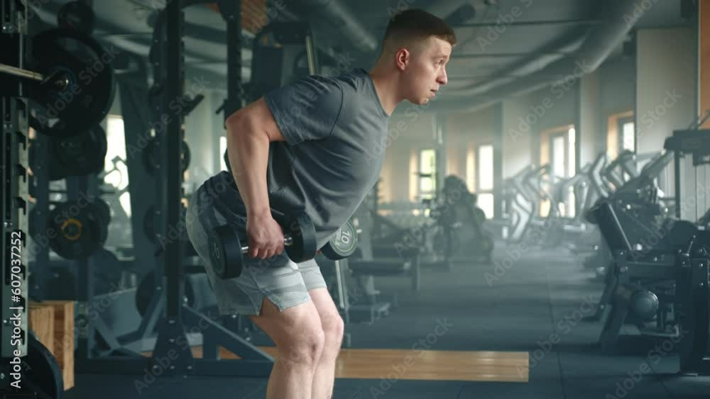 Profile shot of young athlete performing bent over dumbbell row in gym. Sportsman with slightly bent knees leans forward and pulls the weight up to waist. High quality 4k footage Profile shot of young athlete performing bent over dumbbell row in gym. Sportsman with slightly bent knees leans forward and pulls the weight up to waist. High quality 4k footage
