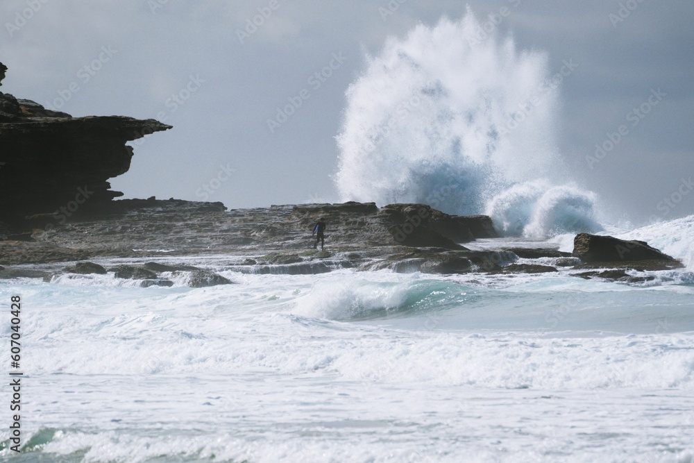 Landscape shot of a surfer standing on the rocky shore of the sea with waves crashing the rocks