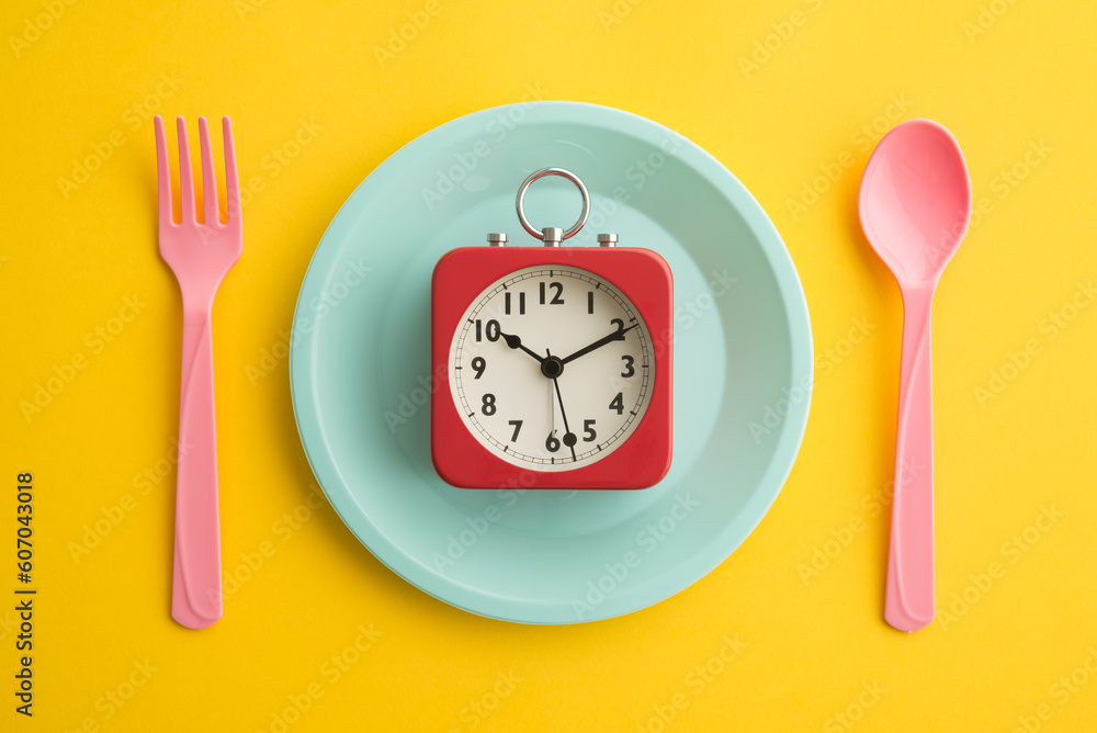 Flat lay of minimal style red retro clock alarm on blue pastel disk, pink fork and spoon on yellow background. IF (intermittent fasting), diet and weight loss, creative idea design, art concept. Flat lay of minimal style red retro clock alarm on blue pastel disk, pink fork and spoon on yellow background. IF (intermittent fasting), diet and weight loss, creative idea design, art concept.