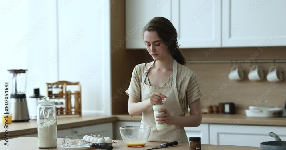 Focused chef girl in baker apron whisking eggs and milk in glass bowl, cooking omelet for breakfast. Positive pretty housewife woman preparing dough for baking pancakes Focused chef girl in baker apron whisking eggs and milk in glass bowl, cooking omelet for breakfast. Positive pretty housewife woman preparing dough for baking pancakes
