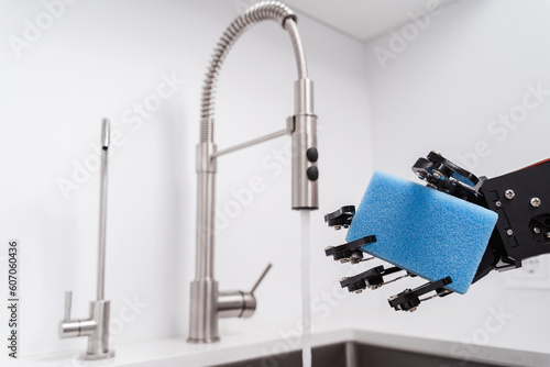 Real robot's hand holding sponge for dishwashing in stainless sink and faucet