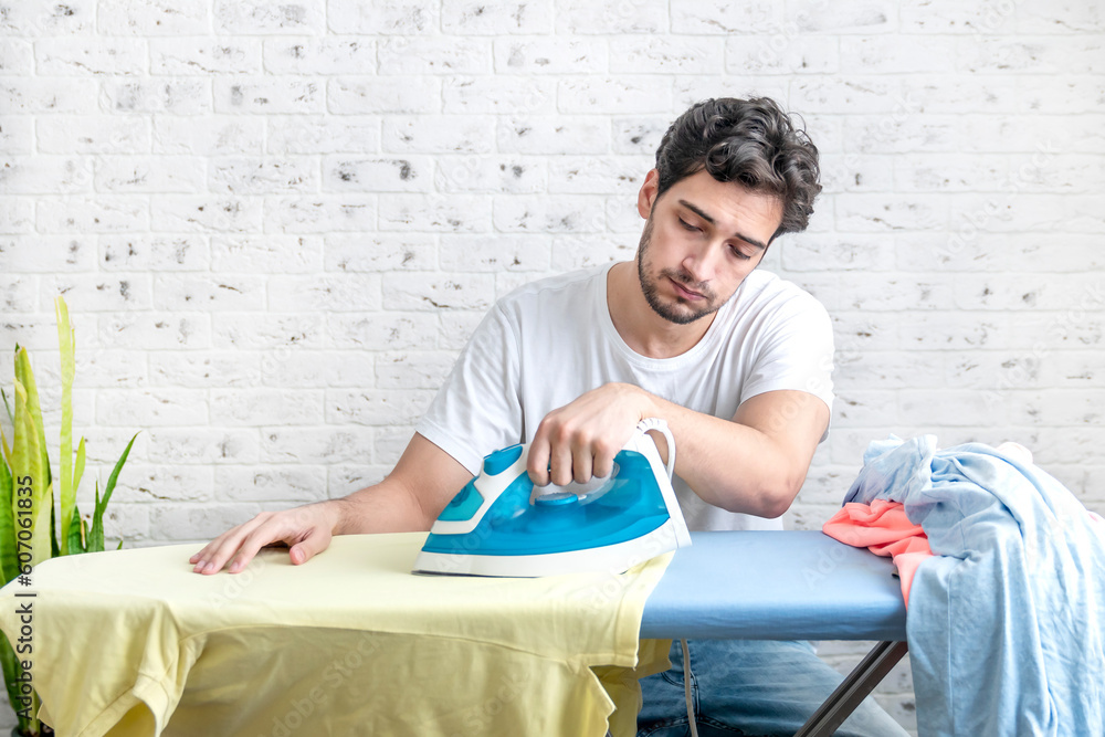 Tired and upset man sitting behind ironing board with many clothes on it. Household and exhausting ironing concept Tired and upset man sitting behind ironing board with many clothes on it. Household and exhausting ironing concept