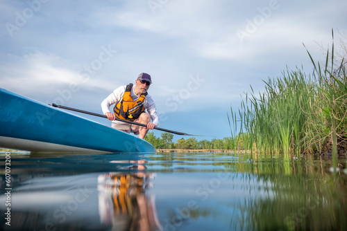 senior male paddler is paddling a stand up paddleboard on a calm lake in early spring, frog perspective from an action camera at water level