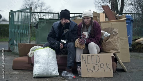 Two dirty and poorly dressed homeless people, a man and a woman, sit by a pile of rubbish with a handwritten NEED WORK poster and eating something. Young woman is sick and coughing.