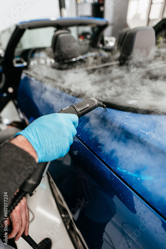 Professional vehicle detailing service in a modern car workshop. Car workshop specialist putting vinyl foil or film on car. Worker using steam pistol for better wrapping and tightening foil edges.