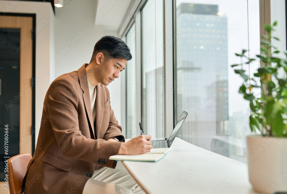 Young busy Asian business man manager working on laptop in office writing notes. Professional businessman analyst using computer checking corporate data, watching online webinar, elearning concept.