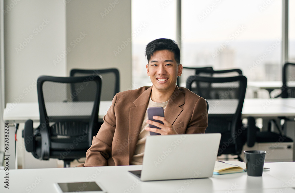 Young happy smiling professional Asian Japanese business man manager executive holding smartphone using mobile cell phone looking at camera working in office with digital cellular technology. Young happy smiling professional Asian Japanese business man manager executive holding smartphone using mobile cell phone looking at camera working in office with digital cellular technology.