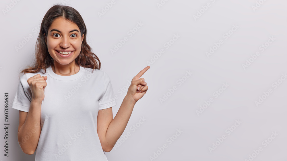 Studio shot of young Iranian woman with dark hair clenches fist points index finger on blank space for your advetising content isolated over white background rejoices good news. Hey check this out Studio shot of young Iranian woman with dark hair clenches fist points index finger on blank space for your advetising content isolated over white background rejoices good news. Hey check this out