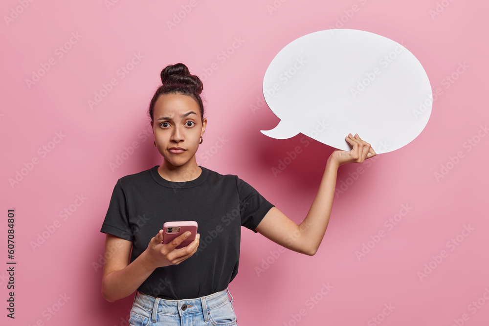 Latin woman holds speech bubble and smartphone invites you to imagine impactful words she may share raises eyebrows poses against pink background. Confused millennial girl with hair bun holds cloud Latin woman holds speech bubble and smartphone invites you to imagine impactful words she may share raises eyebrows poses against pink background. Confused millennial girl with hair bun holds cloud