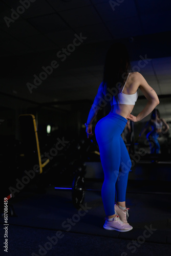 A sports girl in blue leggings and a sexy body warms up in front of a mirror at a workout in a gym with exercise equipment  © Alex May