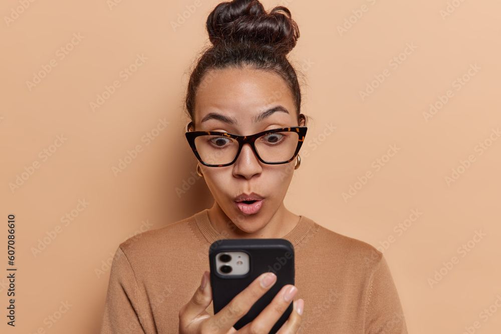Photo of stunned Latin woman stares at smartphone has expression filled with disbelief as she reacts to terrible revelation reads something terrible wears spectacles casual jumper poses indoors - obrazy, fototapety, plakaty 