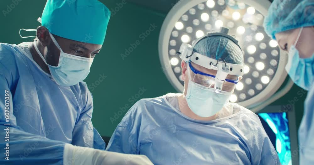 Remote shoot. Three medical workers in operating room. Chief doctor in binoculars. African American assistant in overalls holding scissors. Female intern assists intervention. Doctors during surgery. Remote shoot. Three medical workers in operating room. Chief doctor in binoculars. African American assistant in overalls holding scissors. Female intern assists intervention. Doctors during surgery.
