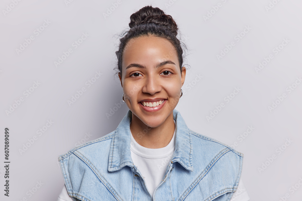 Portrait of pretty good looking Latin woman with dark hair gathered in bun smiles toothily expresses pue happiness brings positive energy to everyone wears denim vest isolated over white background. Portrait of pretty good looking Latin woman with dark hair gathered in bun smiles toothily expresses pue happiness brings positive energy to everyone wears denim vest isolated over white background.