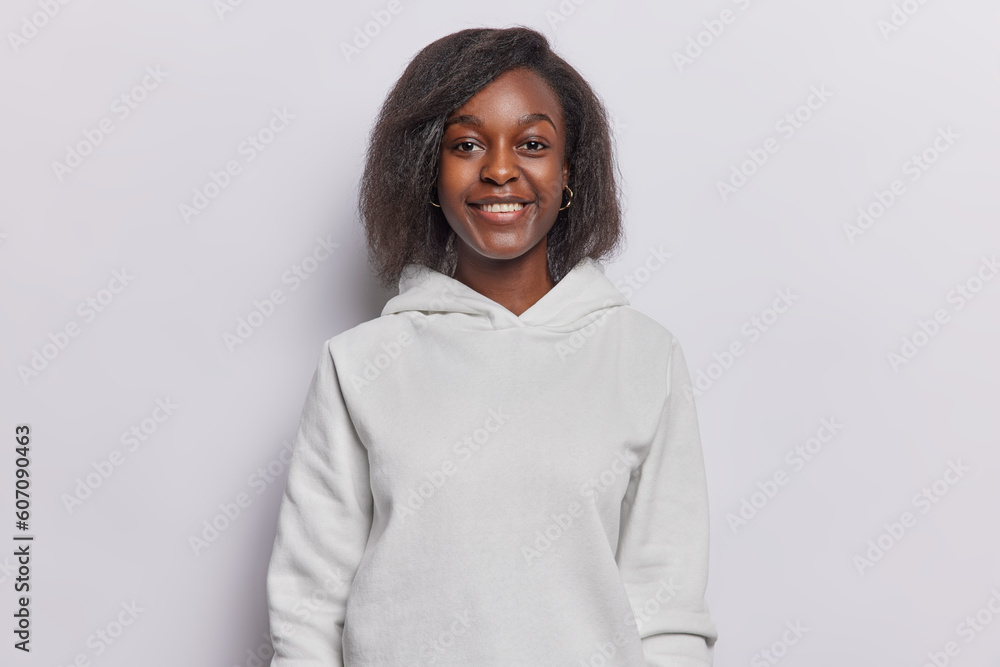Portrait of good looking dark skinned woman with toothy smile dressed in casual sweatshirt looks directly at camera poses against white background. People emotions and face expressions concept Portrait of good looking dark skinned woman with toothy smile dressed in casual sweatshirt looks directly at camera poses against white background. People emotions and face expressions concept