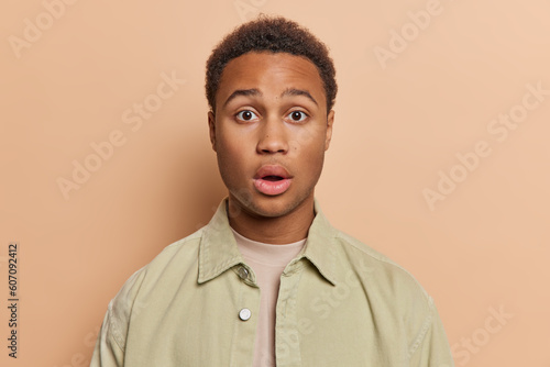 Portrait of handsome young Afro man with short curly hair keeps mouth widely opened holds breath from amazement wears stylish jacket isolated over brown background. People and reactions concept