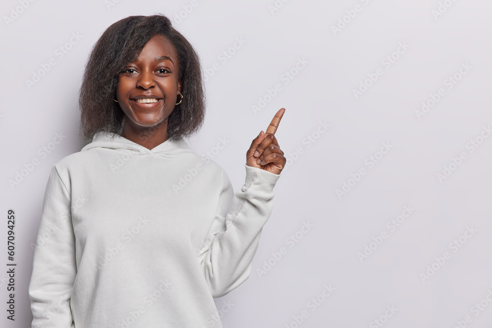 Horizontal shot of attractive dark skinned woman dressed in casual sweatshirt points index finger on blank space advertises product gives direction to something isolated over white background Horizontal shot of attractive dark skinned woman dressed in casual sweatshirt points index finger on blank space advertises product gives direction to something isolated over white background