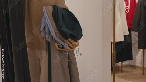 Cropped shot of unrecognizable woman with pile of clothing items on hangers going into fitting room of contemporary designer boutique and closing curtains