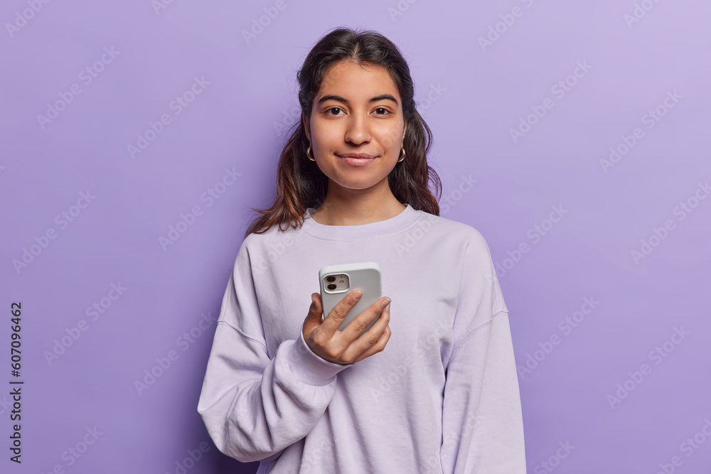 Portrait of pretty brunette woman uses modern phone device checks social network news online reads texting message dressed in pullover isolated over purple background. Ethnicity and technology concept Portrait of pretty brunette woman uses modern phone device checks social network news online reads texting message dressed in pullover isolated over purple background. Ethnicity and technology concept