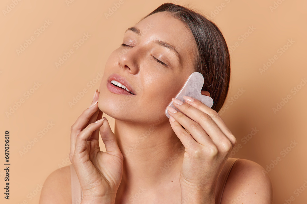 Skin treatment and beauty routine concept. Pleased brunette European woman massages face with jade stone touches cheek gently has eyes closed isolated over brown background enjoys sensation.