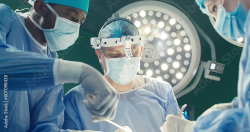 Group of medical workers conducts operation. Doctor in overalls and binoculars holds scalpel. African-American in mask and cap holds scissors. Female intern assist at background of operation lamp.