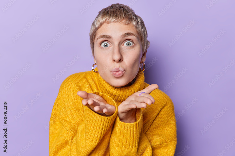 Portrait of romantic short haired young woman keeps lips rounded blows mwah or sweet kiss at camera dressed in comfortable yellow jumper poses against purple background. Moment of pure sweetness Portrait of romantic short haired young woman keeps lips rounded blows mwah or sweet kiss at camera dressed in comfortable yellow jumper poses against purple background. Moment of pure sweetness