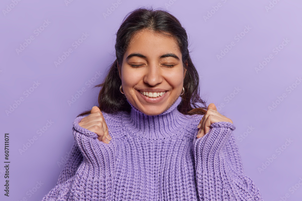 Beautiful cheerful woman from Iran awaits for sommething nice clenches fists raises hands being in anticipation of long awaited meeting smiles gladfully wears knitted jumper isolated over purple wall Beautiful cheerful woman from Iran awaits for sommething nice clenches fists raises hands being in anticipation of long awaited meeting smiles gladfully wears knitted jumper isolated over purple wall
