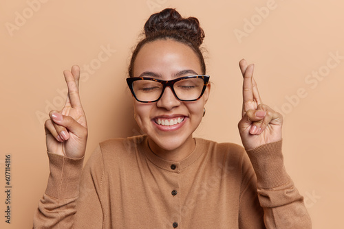 Optimistic woman keeps her fingers crossed hopes for good luck and positive outcomes has desire for success embodies hope wears spectacles and jumper isolated on brown background. Hopeful expectations