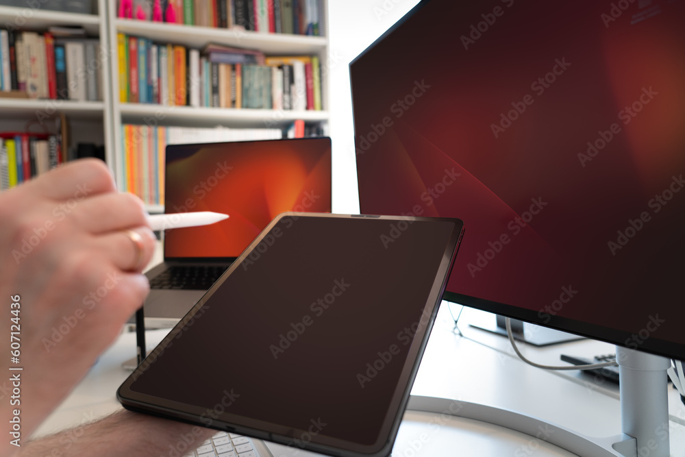 Tablet screen view. Hand holding a graphic pen. Computer and monitor display screens in background. Screens mockup. Home office studio. Tablet screen view. Hand holding a graphic pen. Computer and monitor display screens in background. Screens mockup. Home office studio.