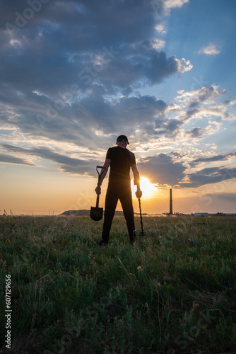 A man in black with a metal detector and a shovel at sunset. Search for jewelry, metal, coins, treasures