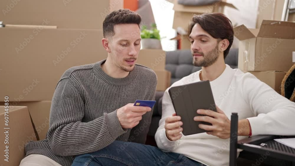 Two men couple shopping with touchpad and credit card sitting on floor at new home Two men couple shopping with touchpad and credit card sitting on floor at new home