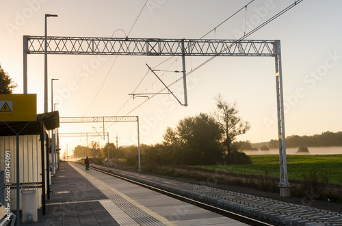 Serenity at Sunset: Railway Landscape with Train Tracks Leading into the Distance, Station Stop, Fields, and a Person Waiting for the Train