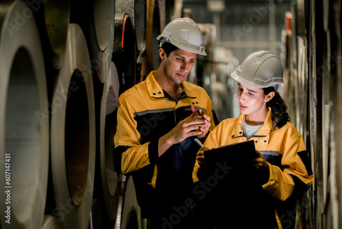 Two maintenance engineers men and women industrial factory behind she talking with workers. They work a heavy industry manufacturing factory. Female industrial engineer wearing a white helmet.