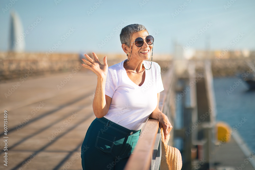 Smiling Mature Woman Waving Hand Posing At Pier With Sailboats