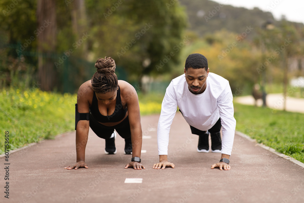 Obraz premium Serious young black couple do push-ups and look at phone, enjoy workout in summer park